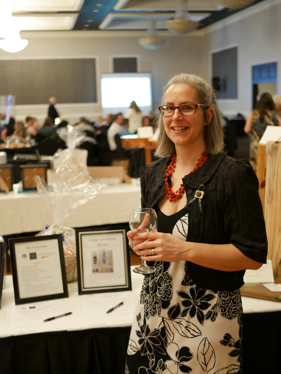 A woman in a black and white dress stands in front of a silent auction table. She holds a glass of wine in her hands.